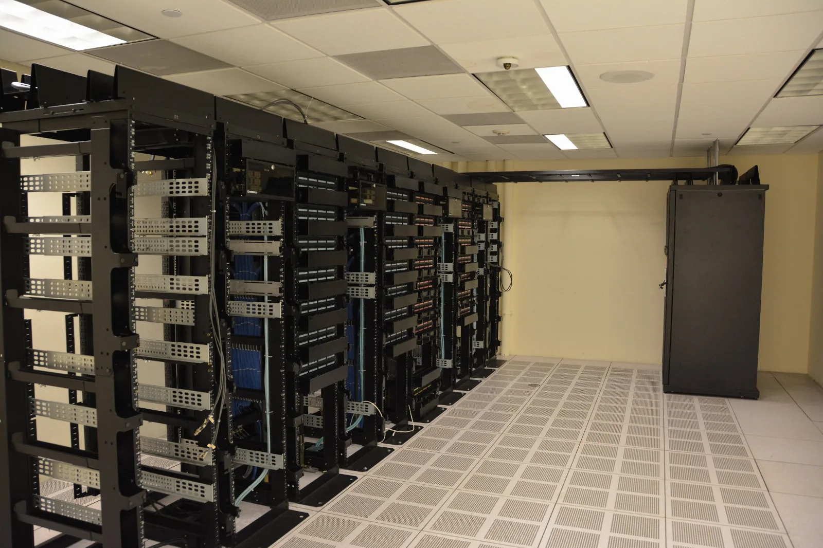 Fileiras de racks de servidores pretos, com cabos azuis, em um data center de piso elevado